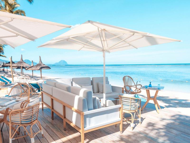 Área de lounge con sofás blancos y sombrillas en la playa bajo un cielo azul.