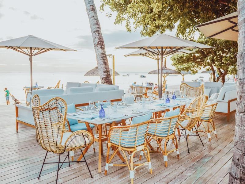 Área exterior luminosa con mesa y sillas en terraza de madera bajo sombrillas, vista al mar.