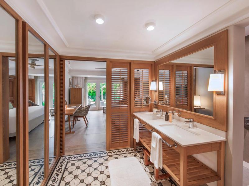 Área moderna de baño de hotel con doble lavabo y muebles de madera junto al acceso al dormitorio.