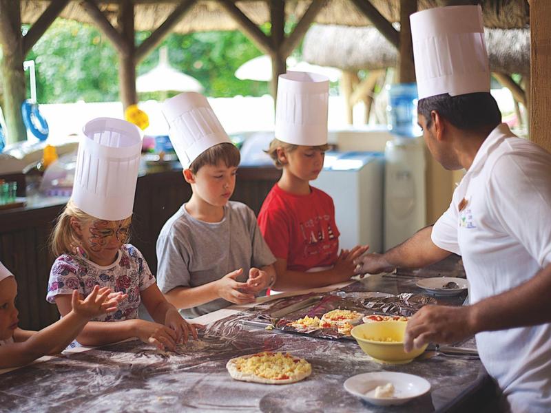 Niños con gorros de chef preparando pizza en una cocina exterior cubierta con un chef.