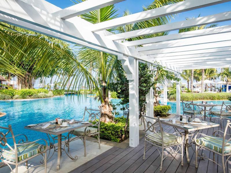 Terraza con mesas y sillas junto a una piscina, rodeada de palmeras y cielo azul.
