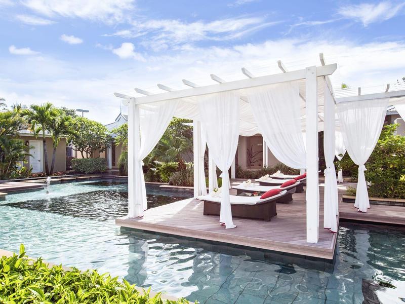 Área de piscina con camas dosel blancas y cojines rojos en un hotel tropical.