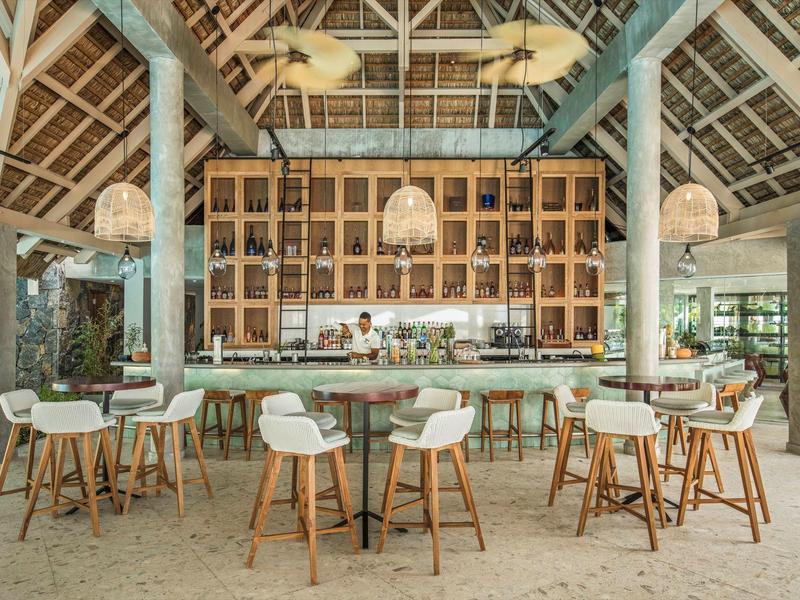 Área de bar moderna en hotel con taburetes de madera y techo abierto de madera y hojas de palma.