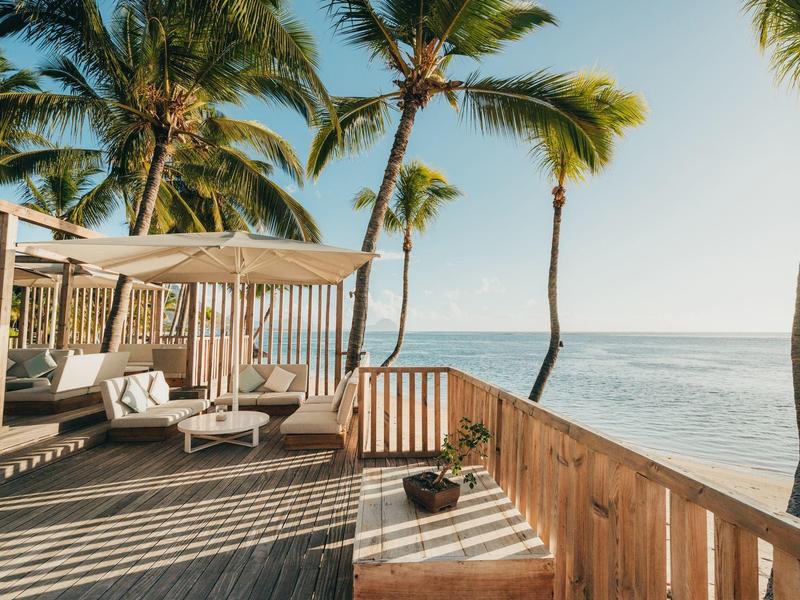 Terraza de madera con tumbonas y palmeras junto al mar bajo cielo despejado.