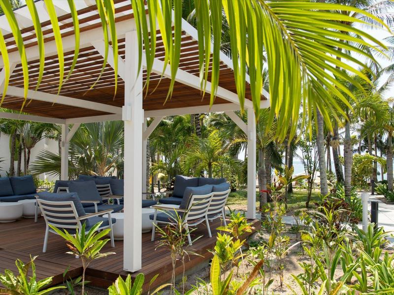 Area lounge esterna con sedie blu sotto un pergolato bianco circondato da piante tropicali.