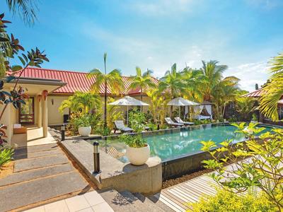 Área de piscina tropical con tumbonas y palmeras frente a un bungalow de techo rojo bajo un cielo azul.
