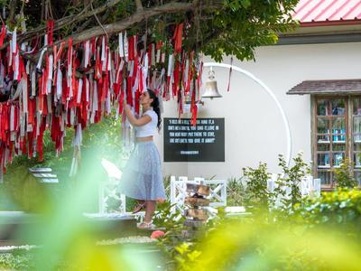 Frau bindet bunte Bänder an Baum, weiße Hauswand und Fenster im Hintergrund, grüne Pflanzen davor.