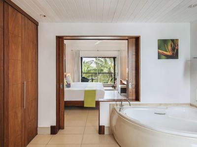 Helles Badezimmer mit großer Badewanne, Holzschrank und Blick in ein Zimmer mit Bett und Fenster.