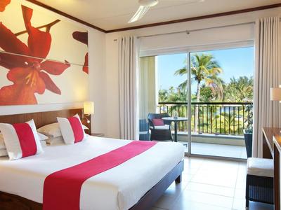 Helles Schlafzimmer mit großem Bett, roter Bettdecke und Balkon mit Blick auf grüne Palmen.