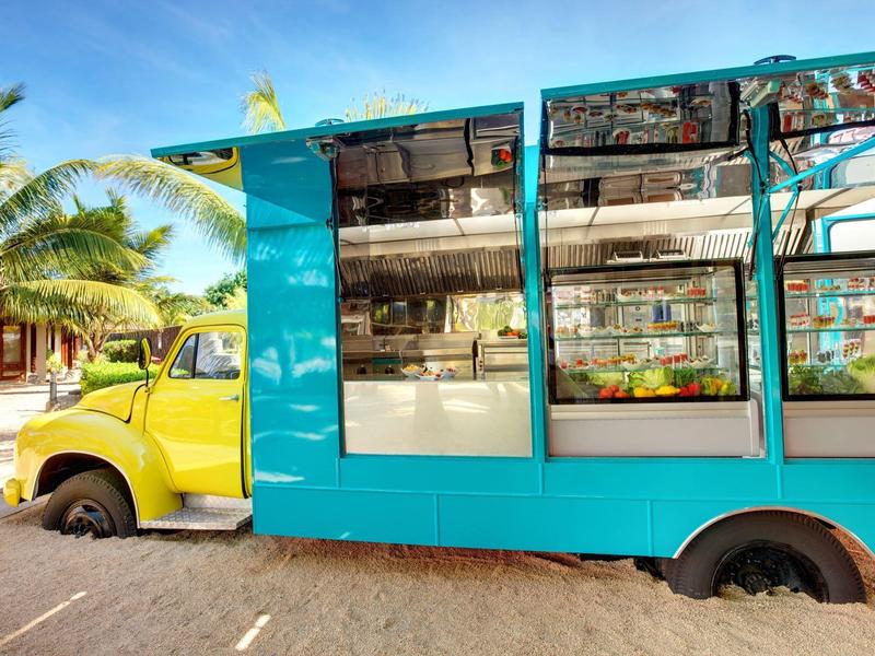 Ein gelbes Fahrzeug mit blauem Anhänger steckt mit Hinterreifen im Sand an einem sonnigen Strand.