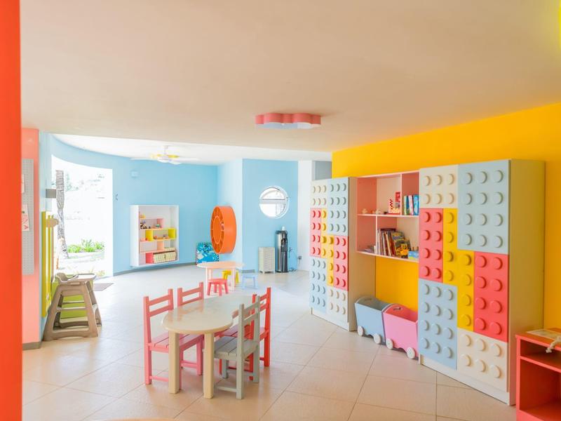 Buntes Kinderspielzimmer mit Lego-Wand, bunten Möbeln, Tischen und Stühlen in hellem Raum.