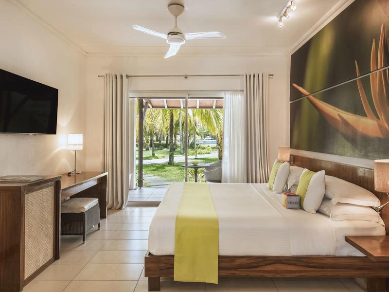 Modernes Schlafzimmer mit großem Bett, Holzmöbeln, TV, Ventilator und Blick auf Garten mit Palmen.