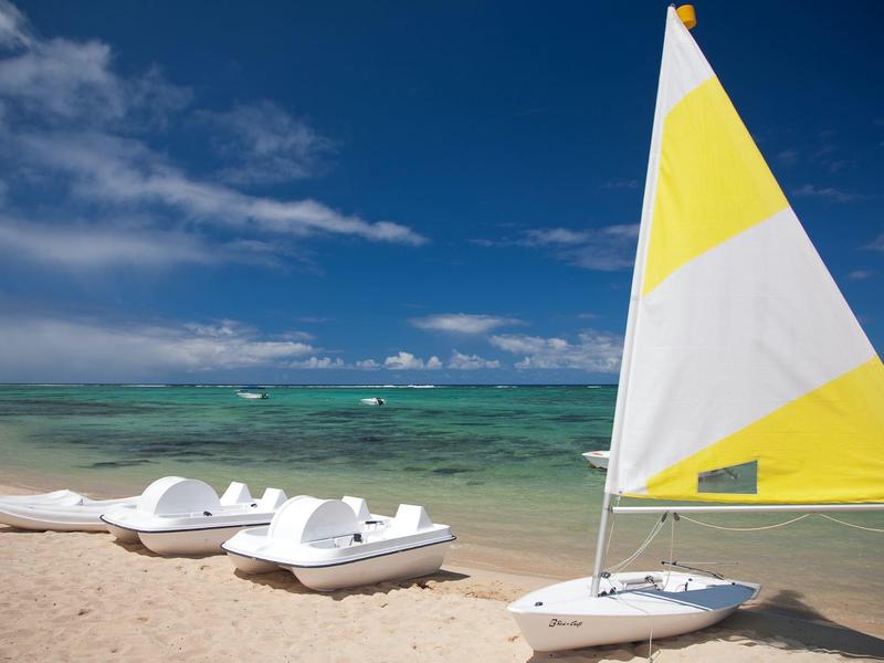 Gelber Segelboot und weiße Tretboote am Sandstrand mit blauem Himmel und grünem Meer.