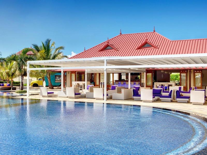 Großer Pool vor Terrasse mit rotem Dach und lila Sitzmöbeln unter blauem Himmel.