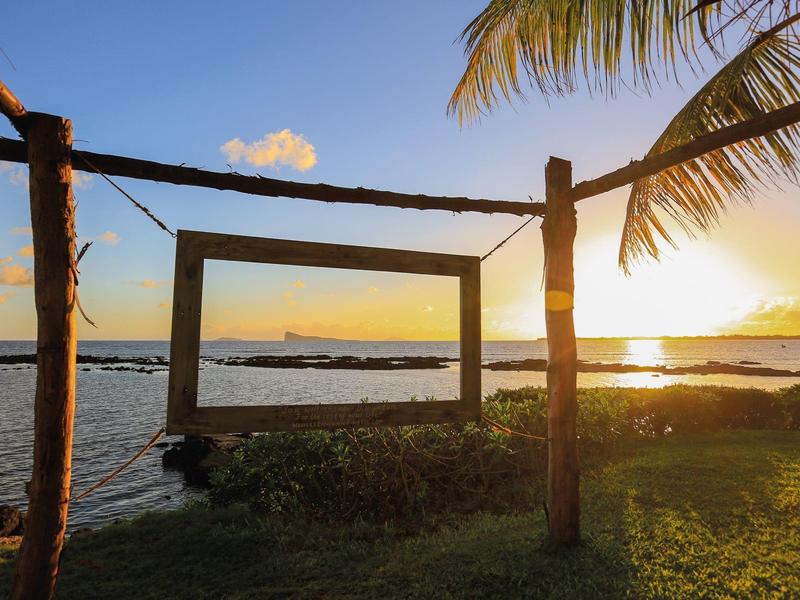 A wooden frame overlooking the coast at sunset, surrounded by palm trees and grass.