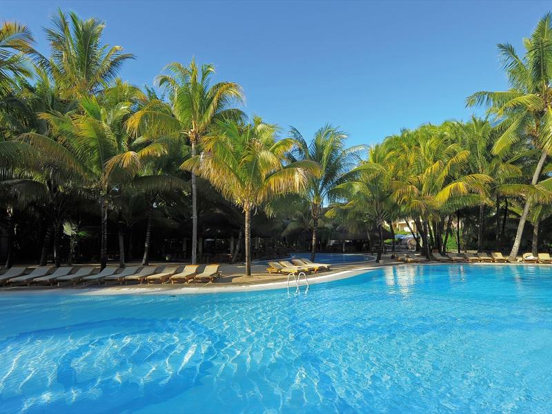 Large clear pool with palm trees and lounge chairs under a bright blue sky.