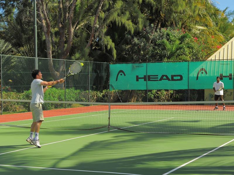 Two men playing tennis on an outdoor green court with palm trees in the background.