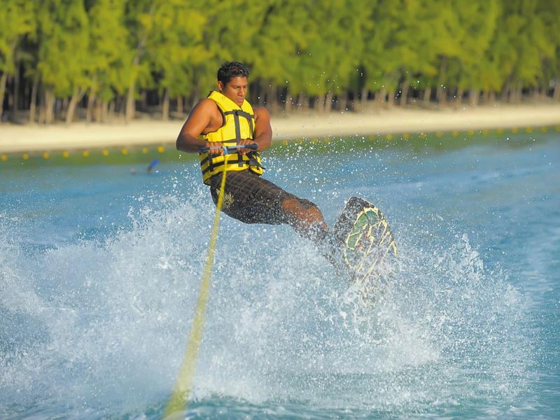 Person wakeboarding on blue water with green shoreline vegetation in the background.