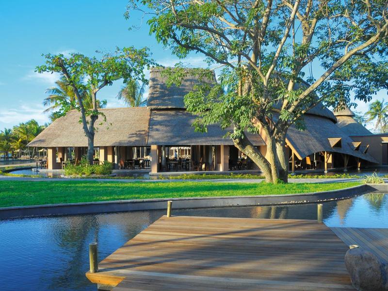 Velký prázdninový dům se slámovou střechou a terasou vedle klidného rybníka pod modrou oblohou.