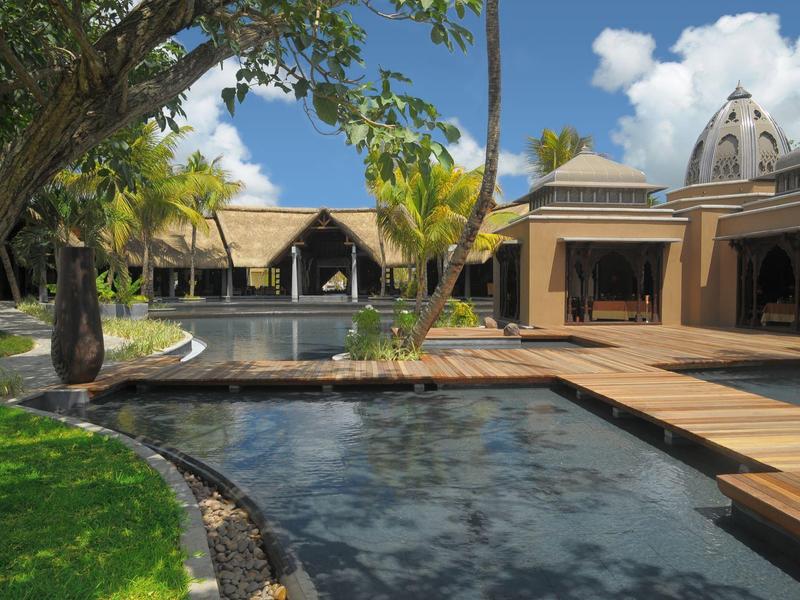 Luxusní hotelový komplex s bazénem, dřevěnými chodníky a tropickými rostlinami pod modrou oblohou.