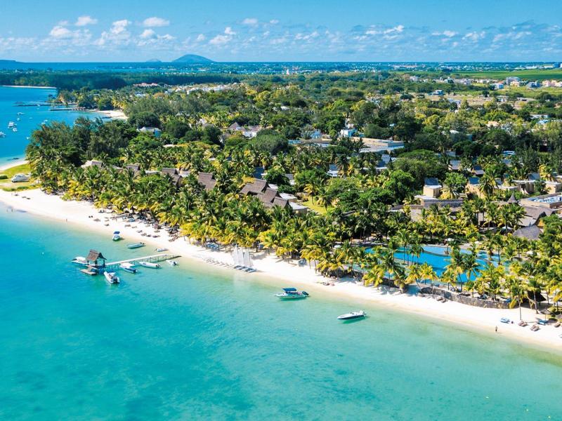 Tropická pláž s čistou vodou, palmami a budovami resortu podél pobřeží.