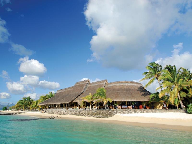 Strand mit türkisfarbenem Wasser, Palmen und einem großen, strohgedeckten Gebäude unter blauem Himmel.