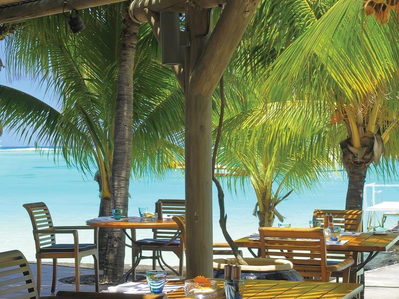 Strandrestaurant mit Holztischen und Stühlen unter Palmen, Blick aufs blaue Meer und sonnigen Himmel.