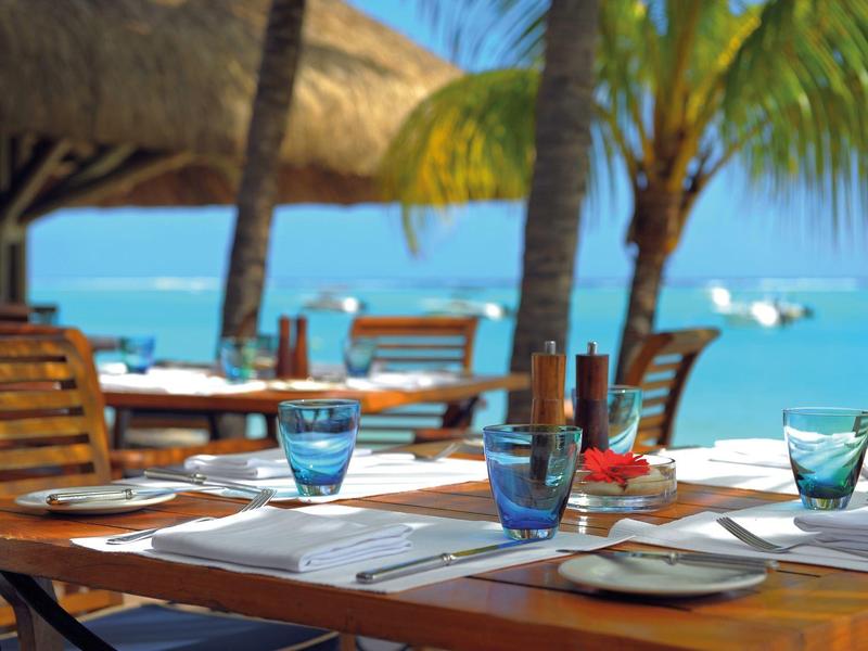 Restaurantterrasse mit Holztischen, blauen Gläsern, Palmen und Blick auf das blaue Meer und Boote.