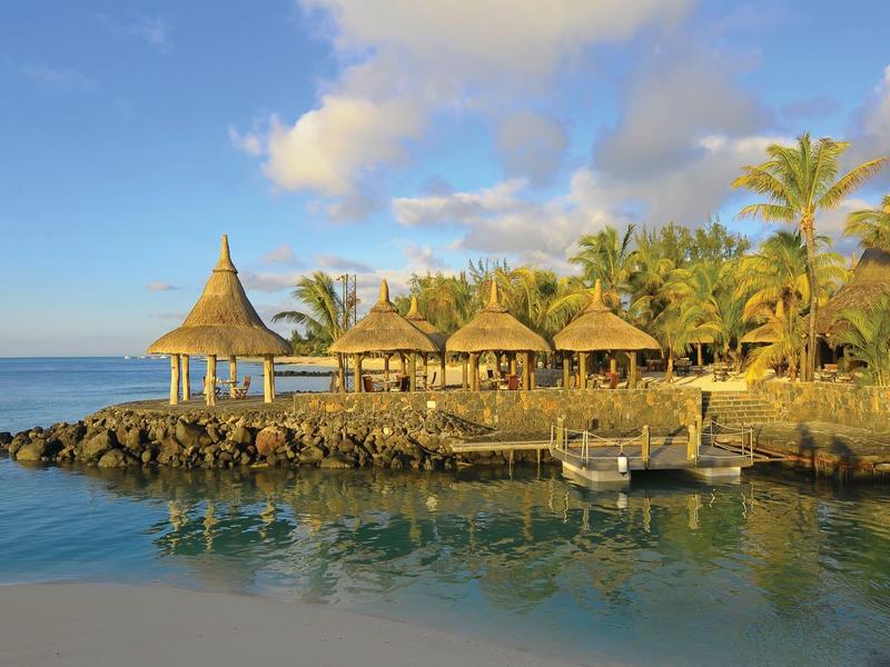 Tropische Strandlandschaft mit Palmen, Strohdächern und klarem blauem Wasser unter blauem Himmel.