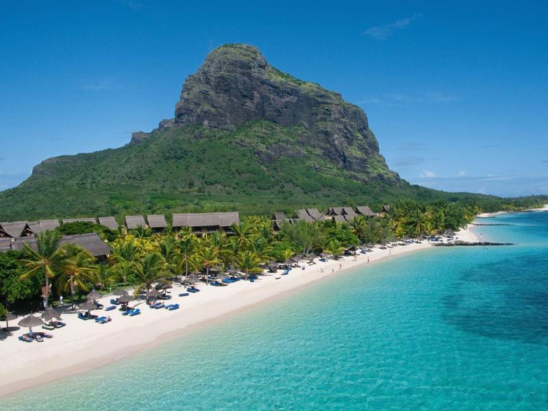 Tropischer Strand mit weißem Sand, Palmen und klarem türkisfarbenem Wasser vor einem Berg.