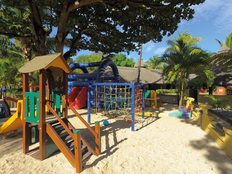 Kinderspielplatz mit Klettergerüst, Rutsche und Sandfläche umgeben von Bäumen und Pflanzen.