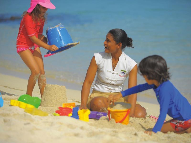 Drei Kinder bauen mit bunten Förmchen am Strand eine Sandburg bei klarem Himmel.