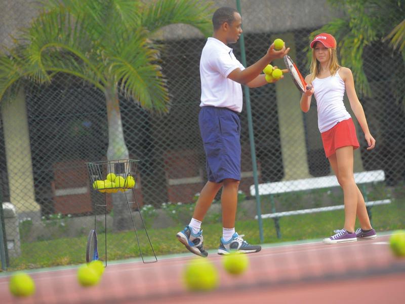 Zwei Personen spielen Tennis auf einem Platz mit Tennisbällen und Palmen im Hintergrund.