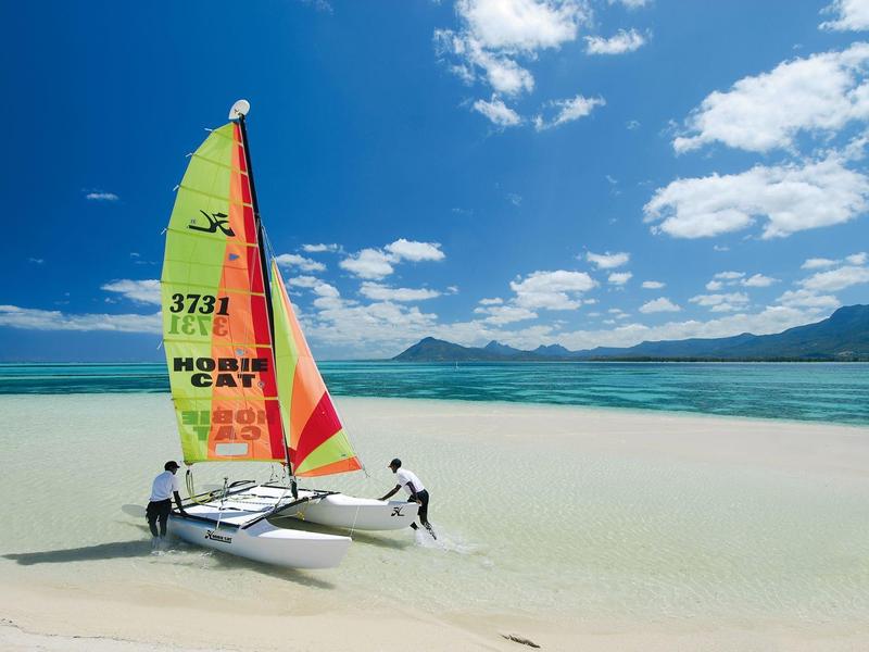 Zwei Personen ziehen ein kleines Segelboot mit buntem Segel durch flaches, klares Wasser an Sandstrand.