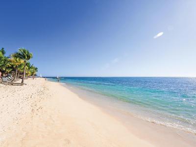 Weißer Sandstrand mit Palmen links, türkisblaues Meer und klarer, blauer Himmel.