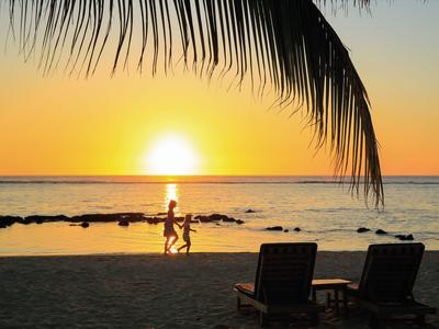 Zwei Personen laufen am Strand bei Sonnenuntergang, Palmenblätter und Liegestühle im Vordergrund.