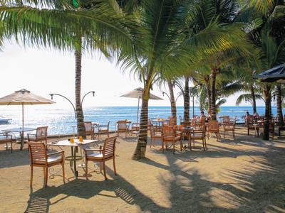 Strandbar mit Holzstühlen und Tischen unter Palmen, Blick auf Meer und Boote bei Sonnenlicht.