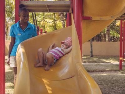 Kind rutscht auf gelber Rutsche, Betreuerin steht daneben im sonnigen Spielplatz.