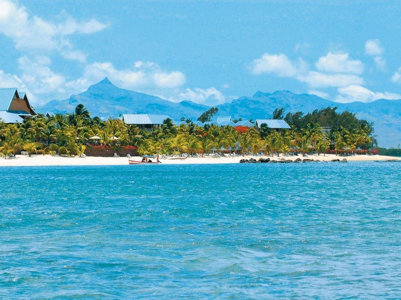 Klarer blauer Himmel über grün bewachsenem Küstenstreifen mit sandigem Strand und Bergen im Hintergrund.