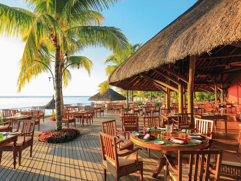Restaurantterrasse mit Holztischen und Stühlen, Palmen, Strohdach und Blick aufs Meer bei Sonnenuntergang.