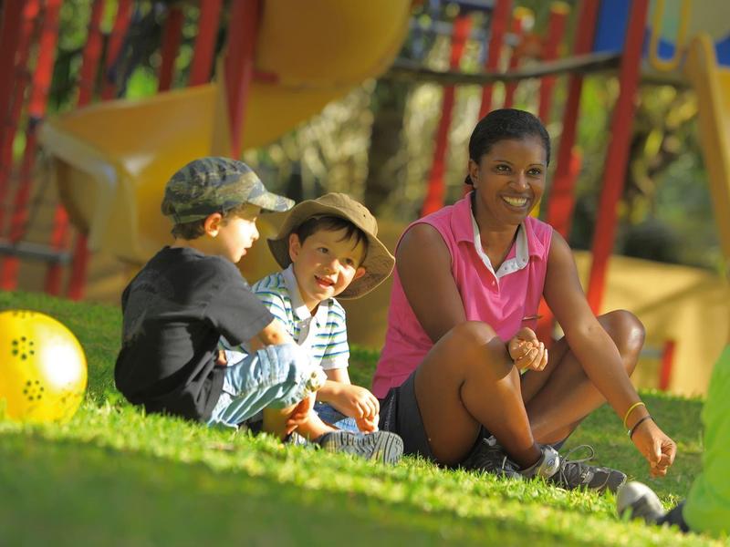 Drei Kinder sitzen auf Gras vor buntem Klettergerüst und Rutsche im sonnigen Park.