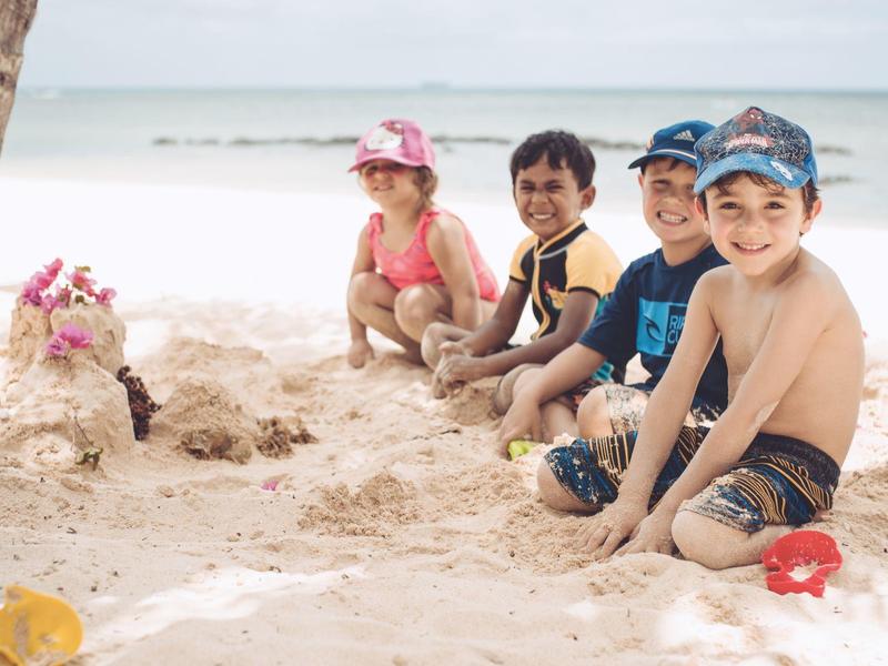 Vier lachende Kinder sitzen am Strand im Sand, im Hintergrund das ruhige Meer unter bewölktem Himmel.