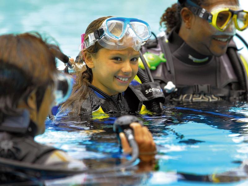 Drei Taucher mit Tauchmasken und Neoprenanzügen im klaren Wasser eines Pools.