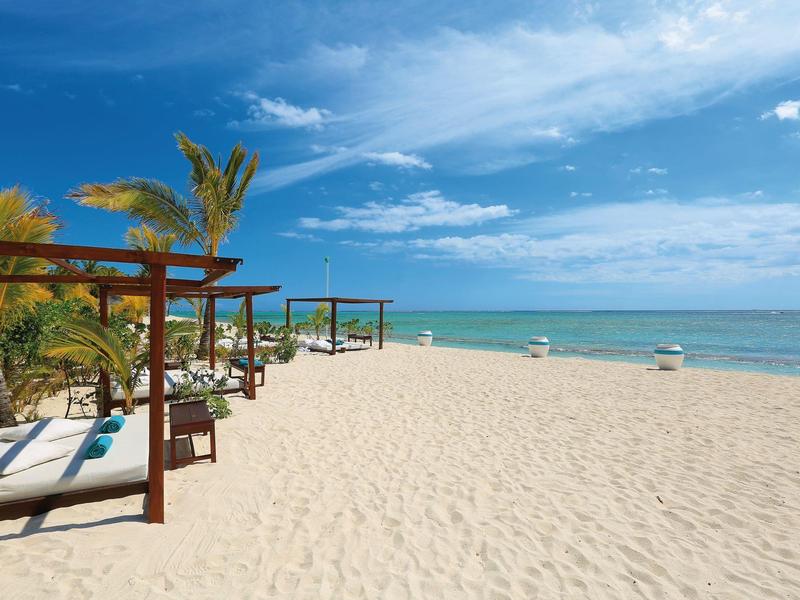 Široká písečná pláž s palmami, dřevěnými lehátky a modrou oblohou u moře