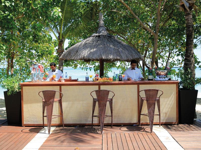 Tři stoličky před plážovým barem se slaměnou střechou a barmany, obklopené stromy.