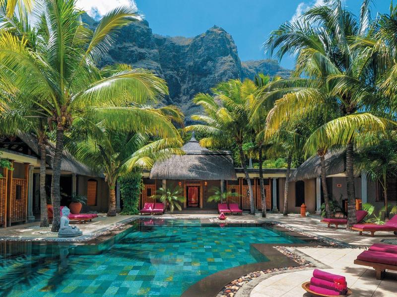 Tropická oblast u bazénu s palmami a červenými lehátky před hotelovou budovou v horách.