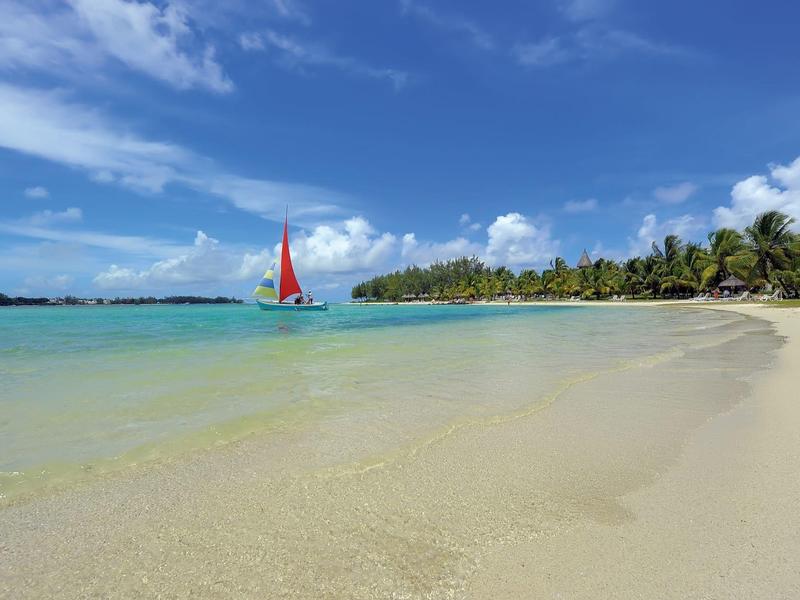 Strand mit klarem Wasser, weißem Sand, Palmen im Hintergrund und einem Segelboot mit rotem Segel.