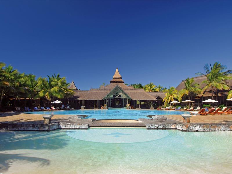 Großer Pool mit Brücke, Palmen und Sonnenschirmen vor einem Gebäude mit spitzem Dach.