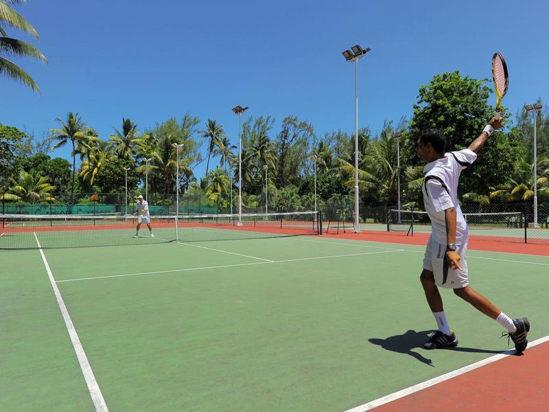 Zwei Männer spielen Tennis auf einem sonnigen, grünen Tennisplatz mit Palmen im Hintergrund.