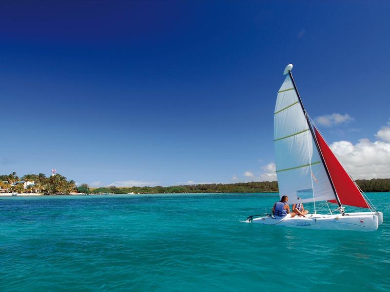 Ein Katamaran mit buntem Segel fährt auf klarem, türkisfarbenem Meer unter blauem Himmel.